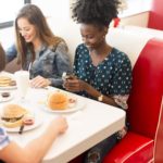 Three young adult friends sit around a vintage restaurant booth eating hamburgers. image represents restaurant stocks to buy